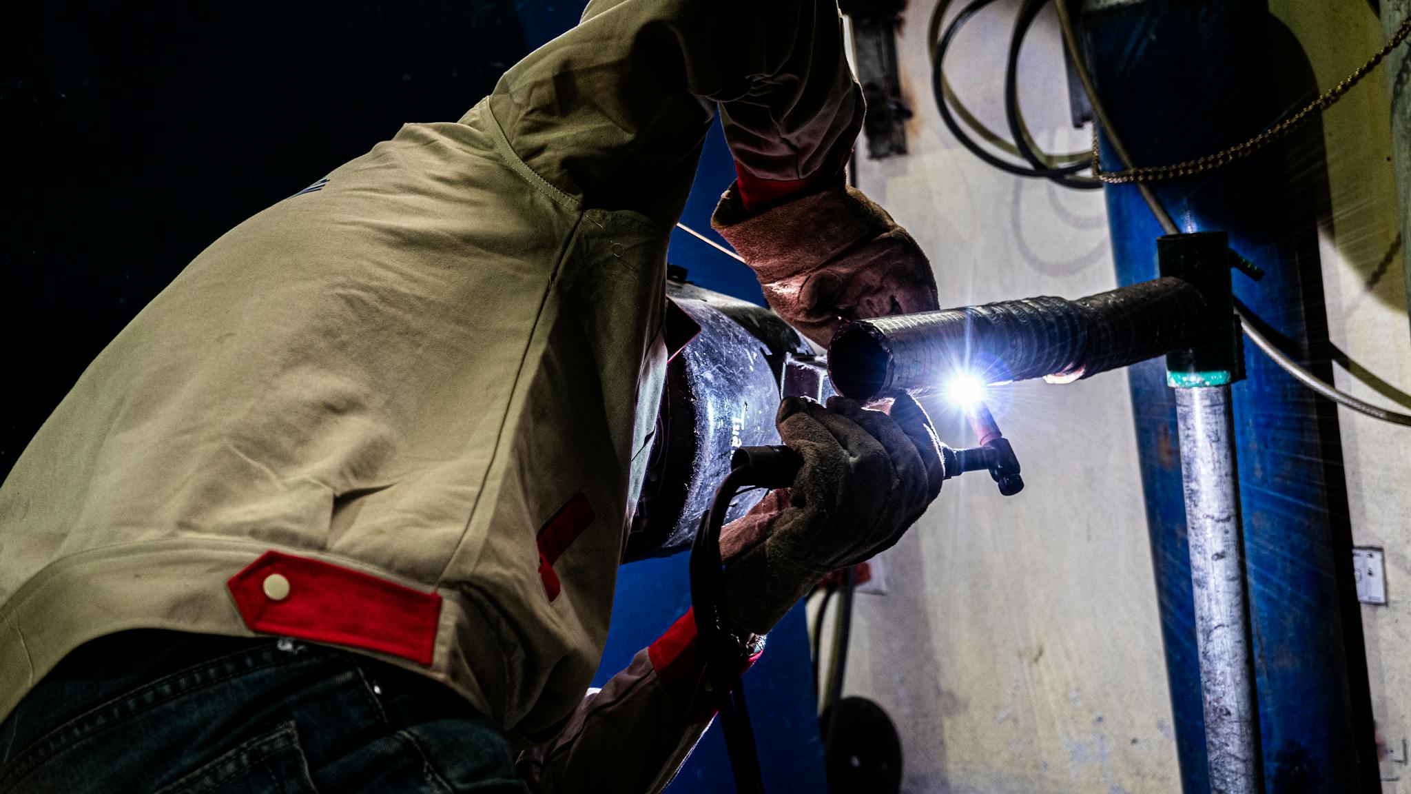 Close-up of a welder working on metal pipes indoors, with bright sparks illuminating the scene.