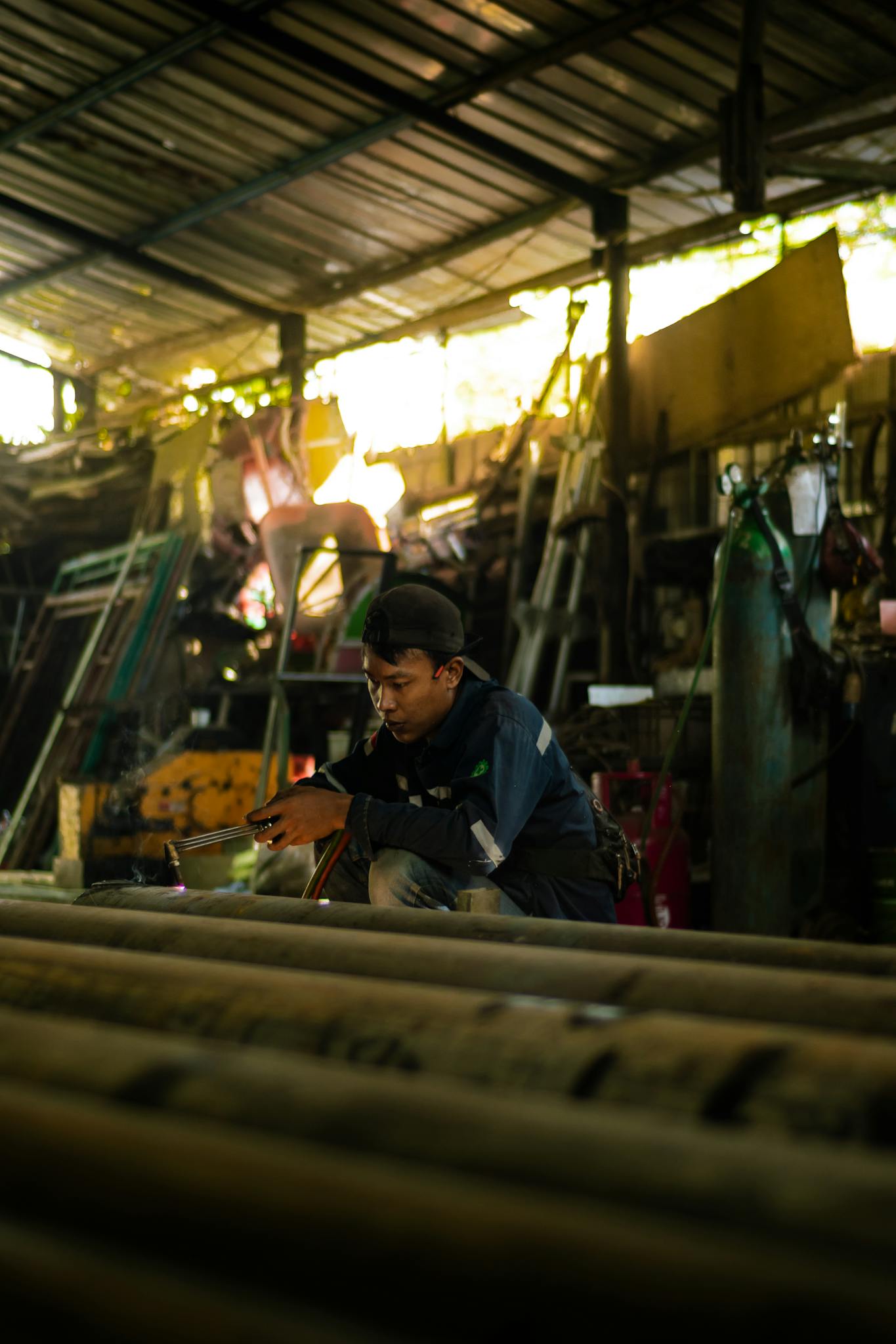 Man welding metal pipes in a cluttered workshop with industrial tools and materials.